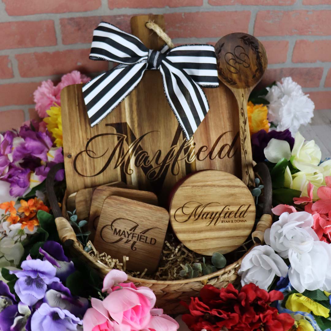 Wooden cutting board and utensils with 'Mayfield' engraving, tied with black-and-white bow, surrounded by colorful flowers.