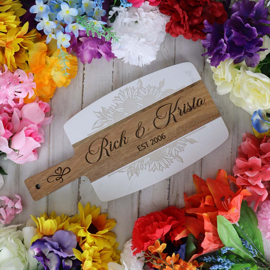 Personalized acacia wood and white marble charcuterie board with 'Rick & Krista EST 2006' engraved, surrounded by colorful flowers.