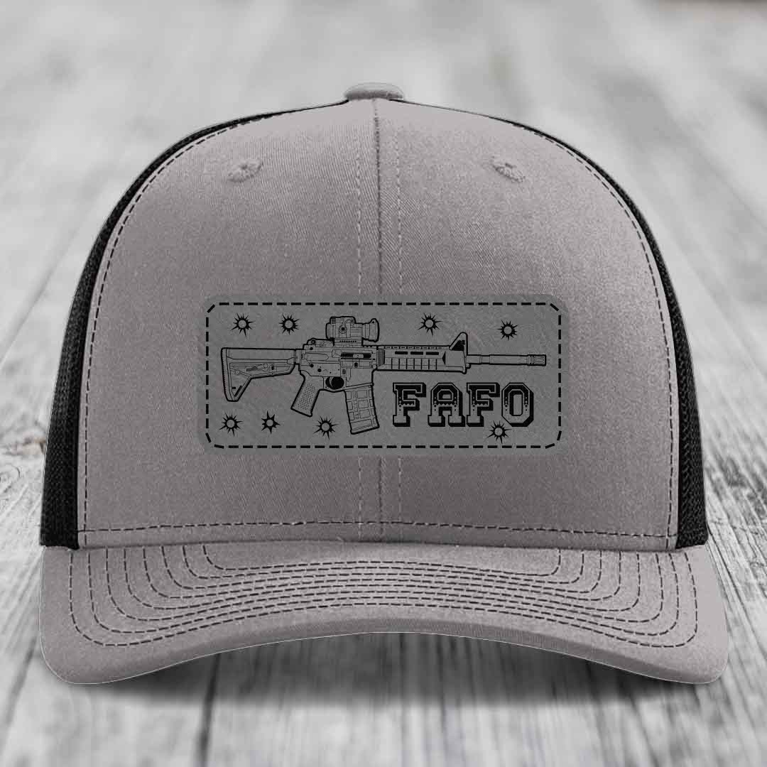 Grey trucker hat with black mesh back and a rectangular leather patch featuring an AR-15 rifle graphic and &