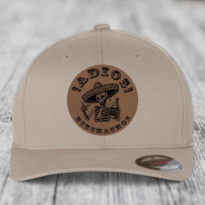 Beige baseball cap with brown circular leather patch featuring skeleton in sombrero holding beer bottle and text &