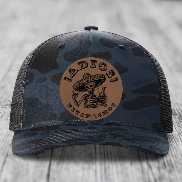 Camouflage trucker hat with brown leather patch featuring skeleton in sombrero holding beer bottle.