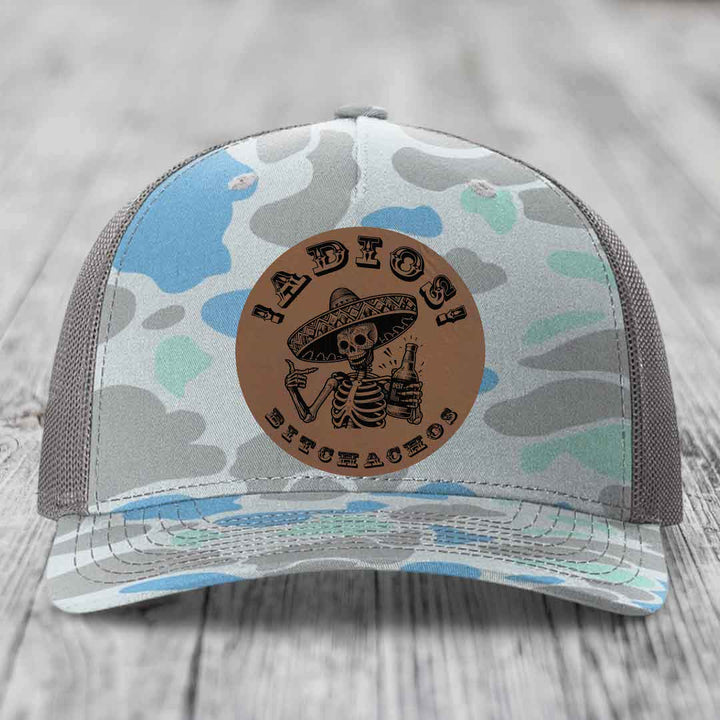 Camouflage trucker hat with brown leather patch featuring skeleton in sombrero holding beer bottle.