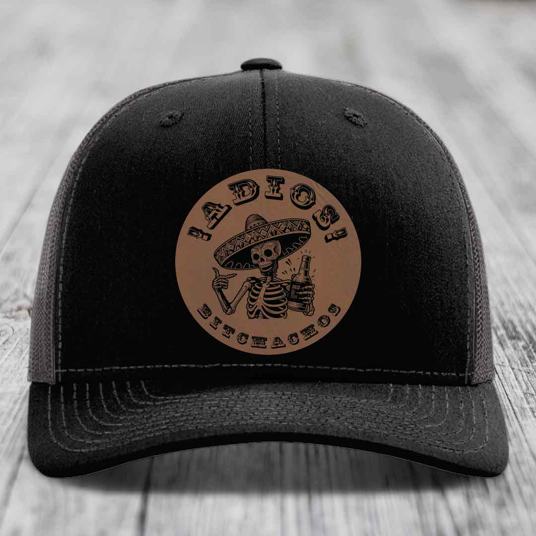 Black trucker hat with brown circular leather patch featuring skeleton in sombrero holding beer bottle.