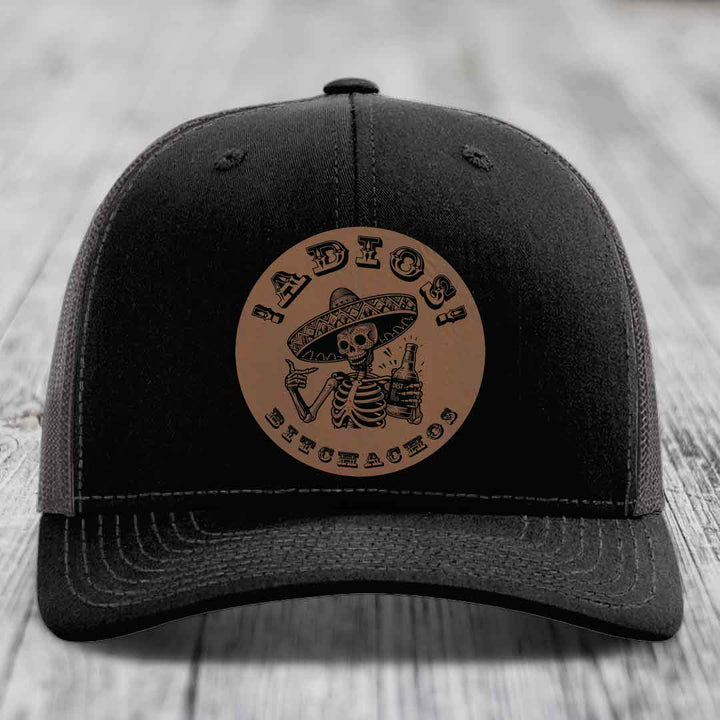 Black trucker hat with brown circular leather patch featuring skeleton in sombrero holding beer bottle.