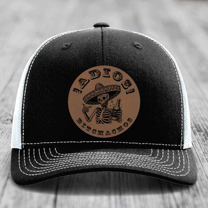 Black trucker hat with white stitching and a brown leather patch featuring a skeleton in a sombrero holding a beer bottle.