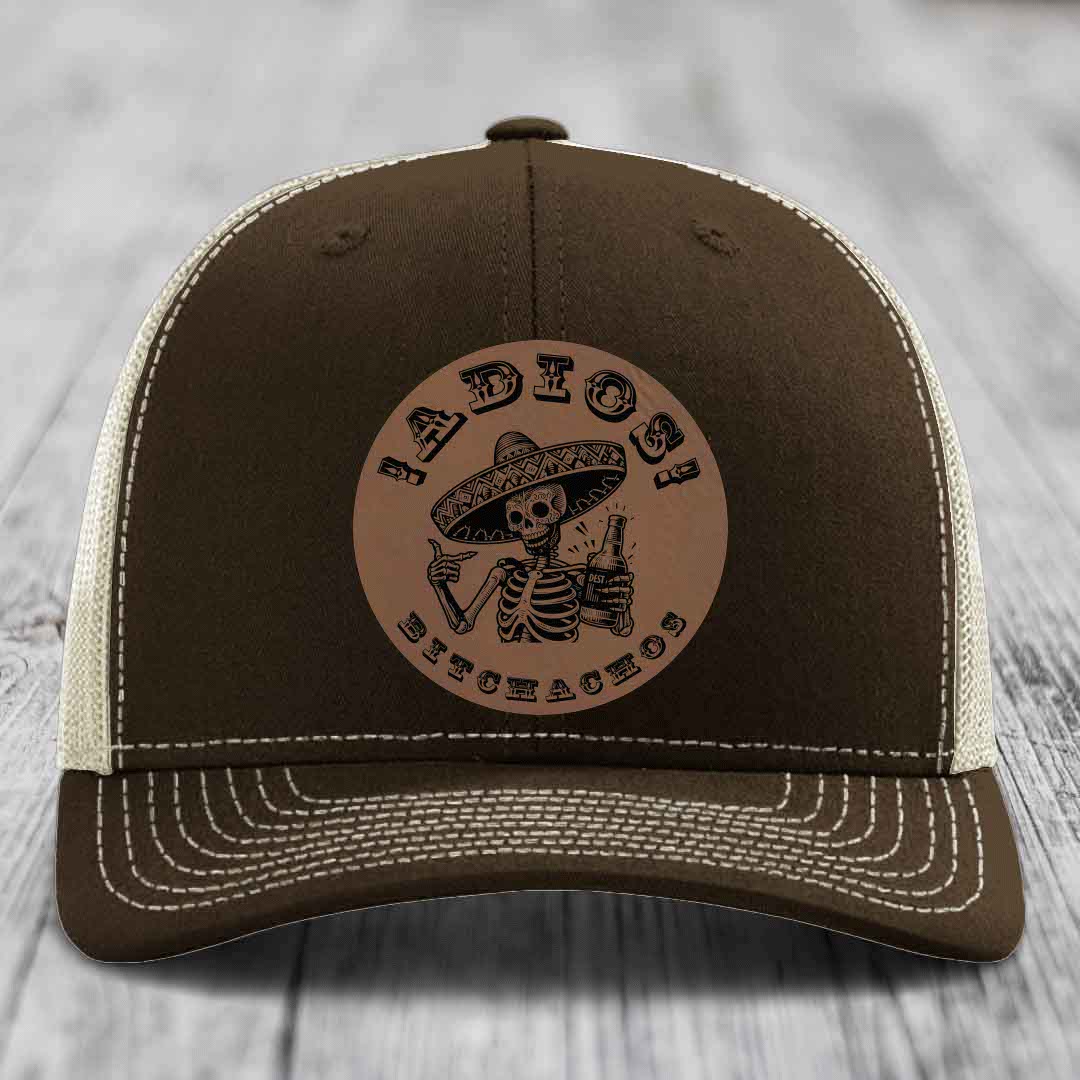 Brown trucker hat with beige mesh back and white stitching, featuring a circular leather patch with a skeleton in a sombrero holding a beer bottle and the text &