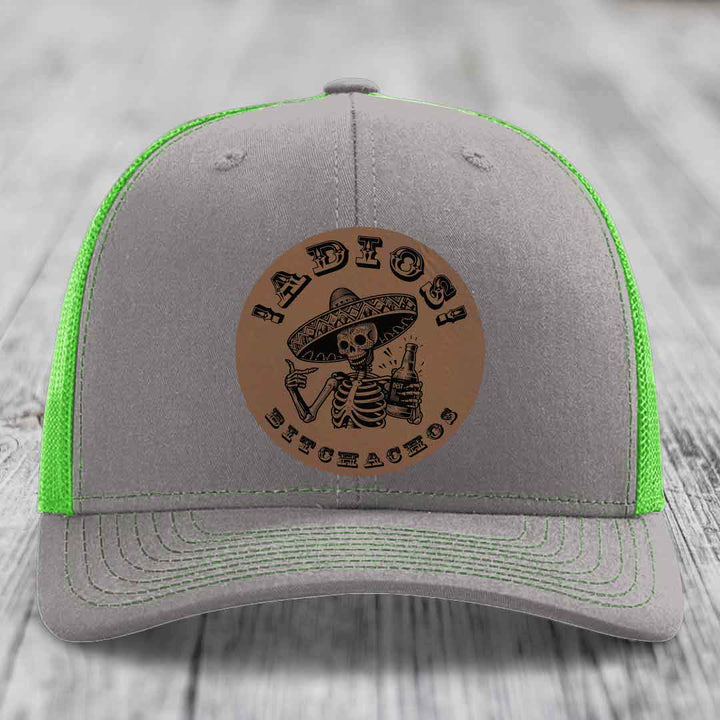 Grey trucker hat with neon green mesh and stitching, featuring a brown leather patch of a skeleton in a sombrero holding a beer bottle with &
