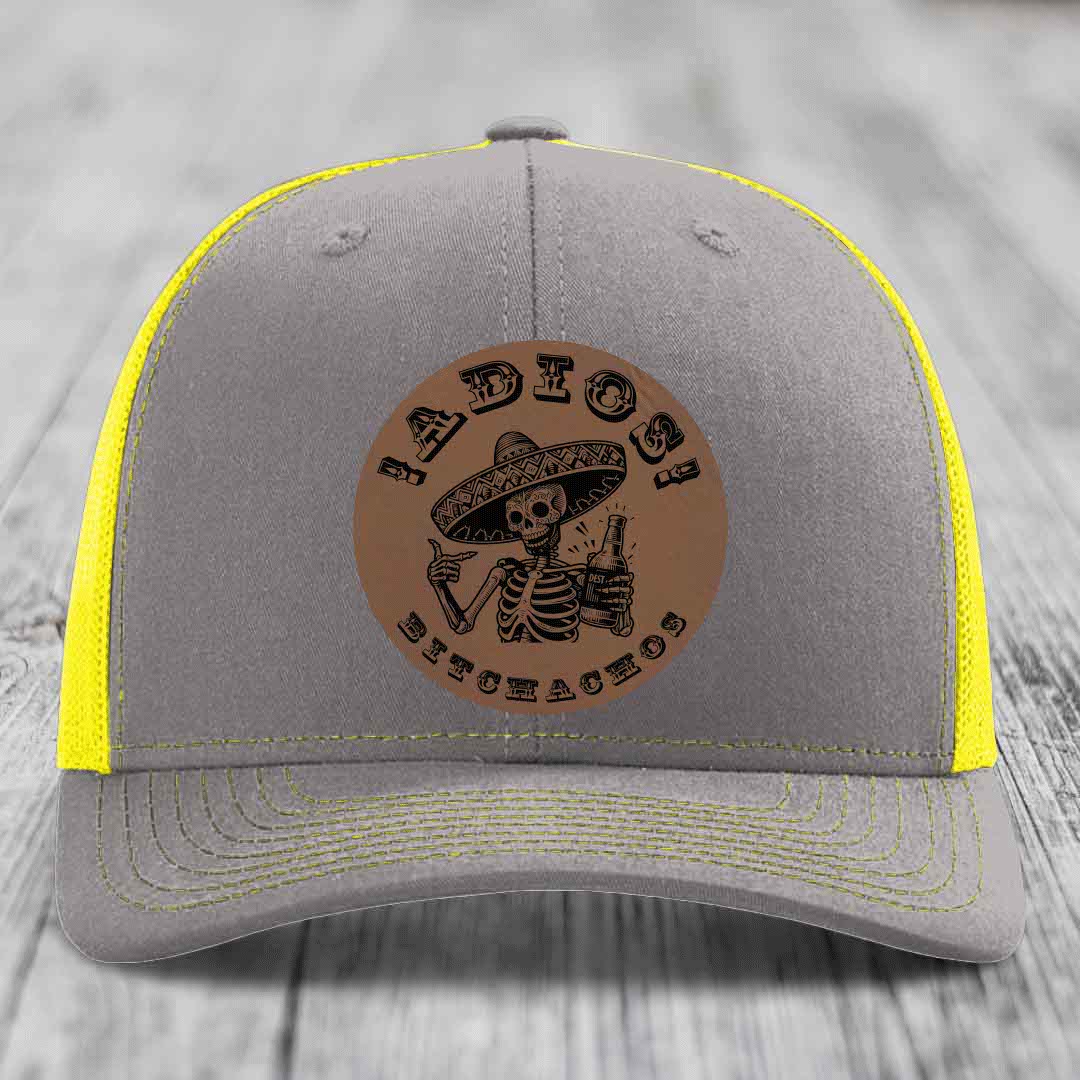 Grey trucker hat with yellow mesh and stitching, featuring a brown leatherette patch of a skeleton in a sombrero holding a bottle with &