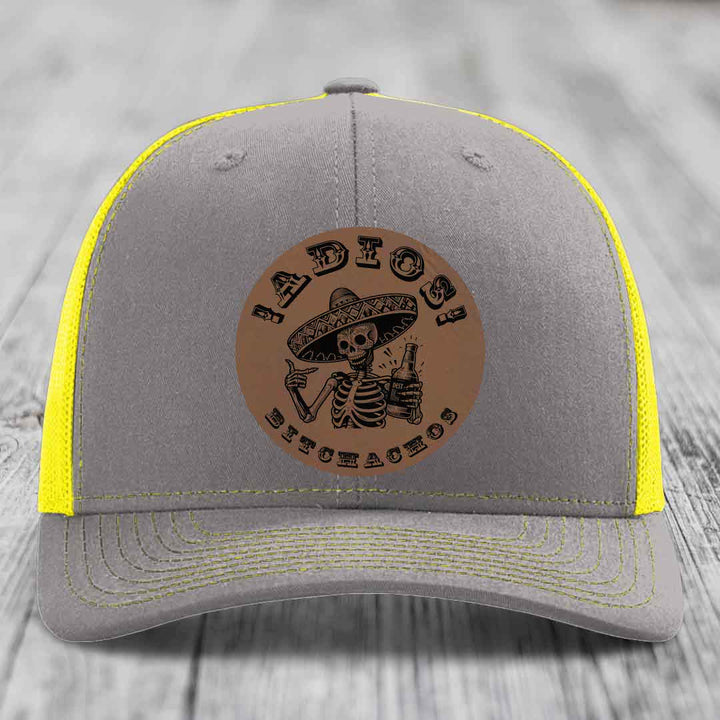 Grey trucker hat with yellow mesh and stitching, featuring a brown leatherette patch of a skeleton in a sombrero holding a bottle with &