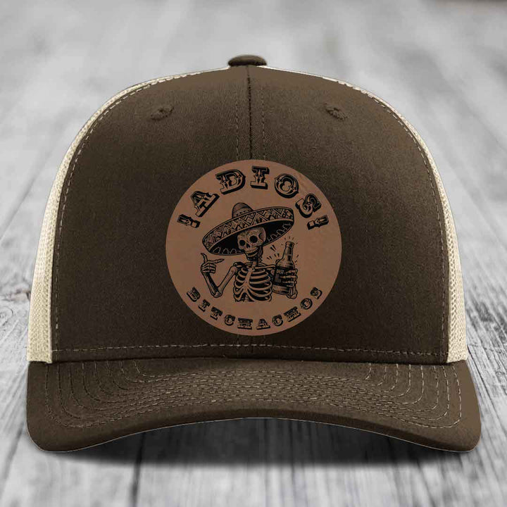 Brown trucker hat with beige mesh back and a circular leather patch featuring a skeleton in a sombrero holding a beer bottle.
