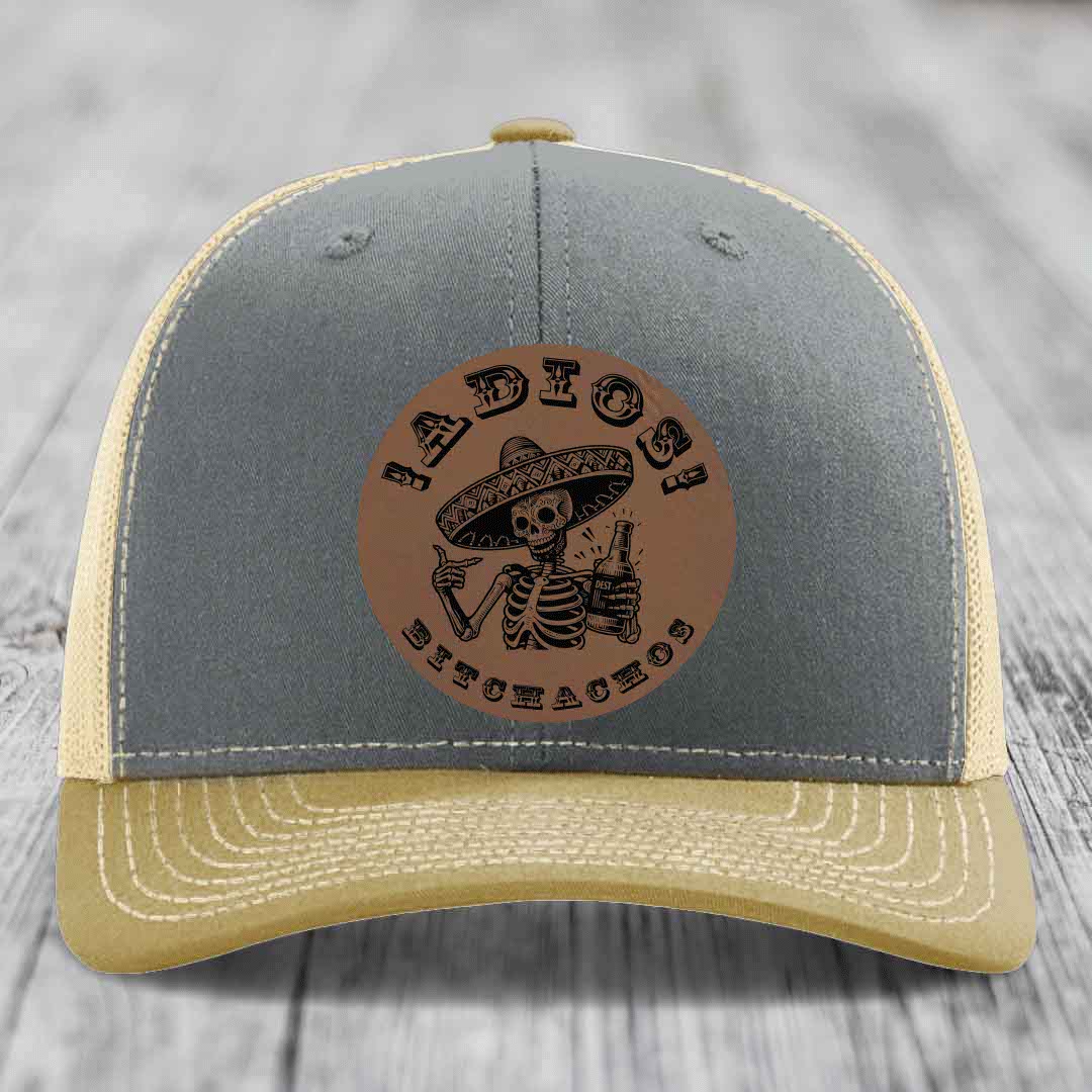 Grey and tan trucker hat with brown leather patch featuring skeleton in sombrero holding beer bottle.
