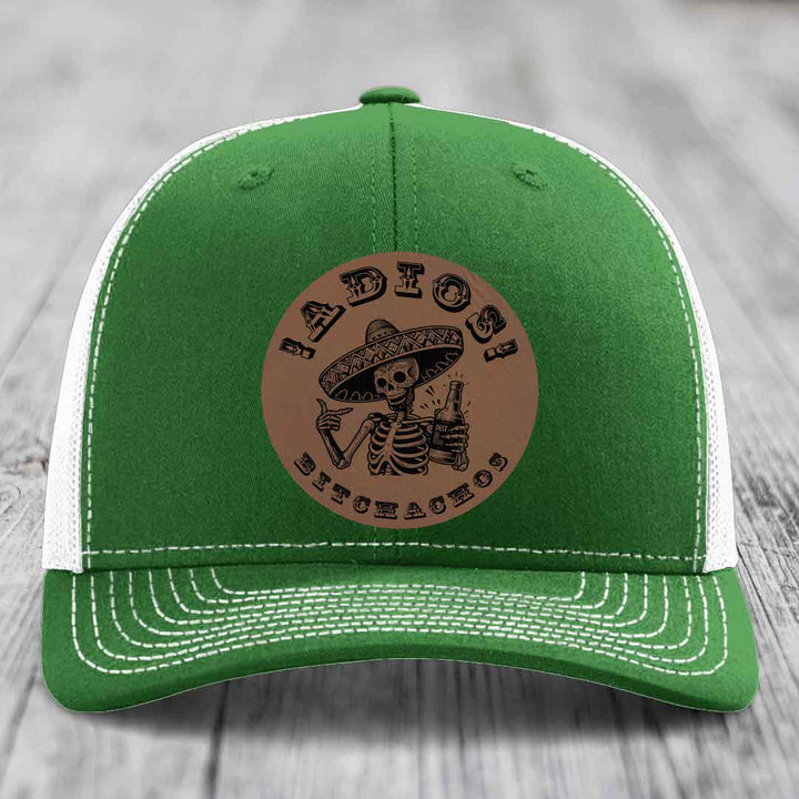 Green trucker hat with white mesh back and stitching, featuring a brown leatherette patch of a skeleton in a sombrero holding a beer bottle with "Adios Bitchachos" text.