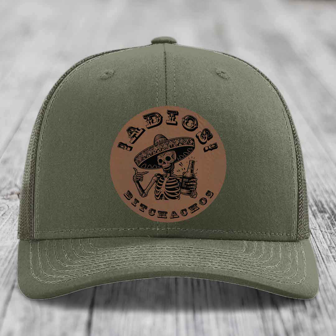 Olive green trucker hat with brown circular leather patch featuring skeleton in sombrero holding beer bottle and text &