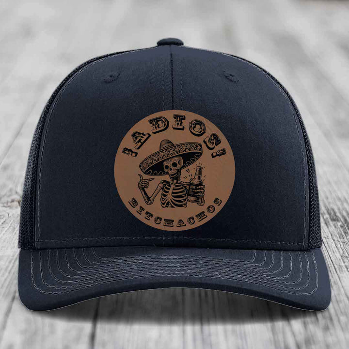 Black trucker hat with brown leather patch featuring skeleton in sombrero holding beer bottle.