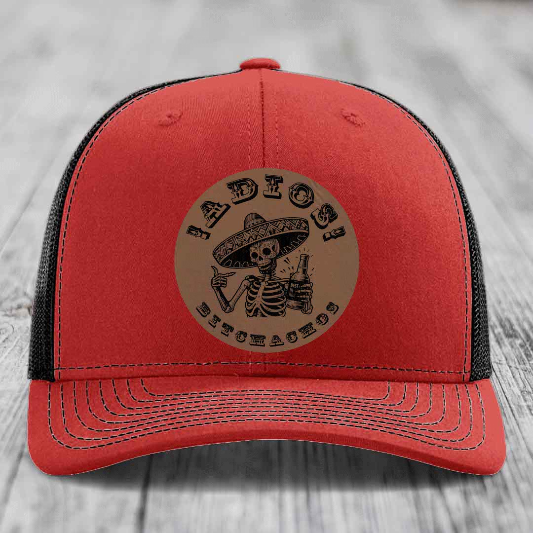 Red trucker hat with black mesh back and a circular tan leather patch featuring a skeleton in a sombrero holding a beer bottle.