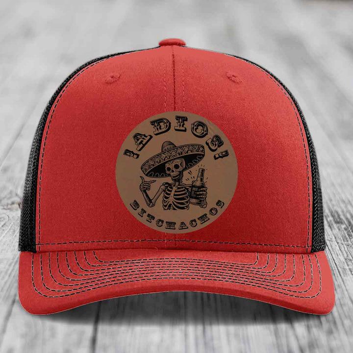 Red trucker hat with black mesh back and a circular tan leather patch featuring a skeleton in a sombrero holding a beer bottle.