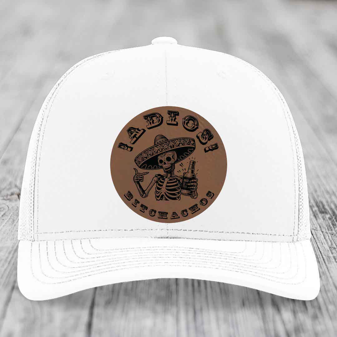 White trucker hat with brown circular leather patch featuring skeleton in sombrero holding beer bottle and text &