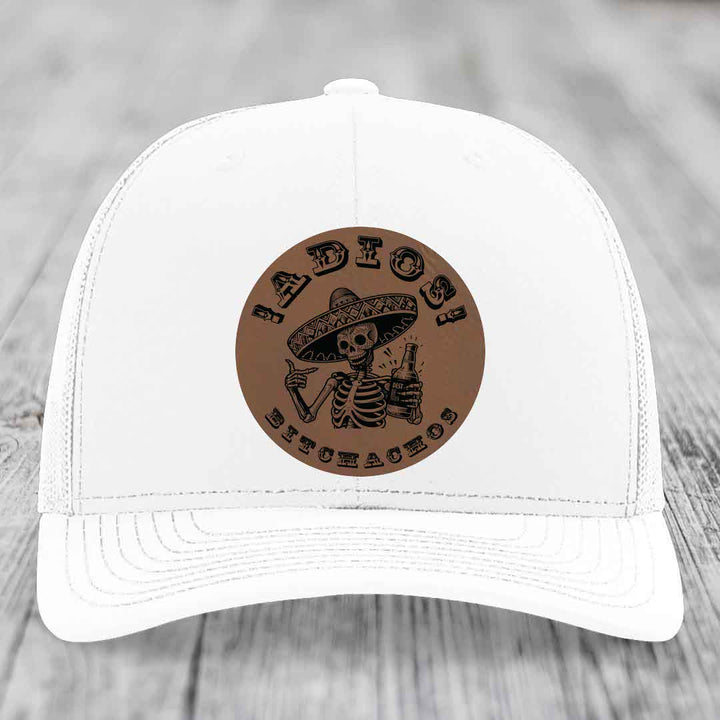 White trucker hat with brown circular leather patch featuring skeleton in sombrero holding beer bottle and text &