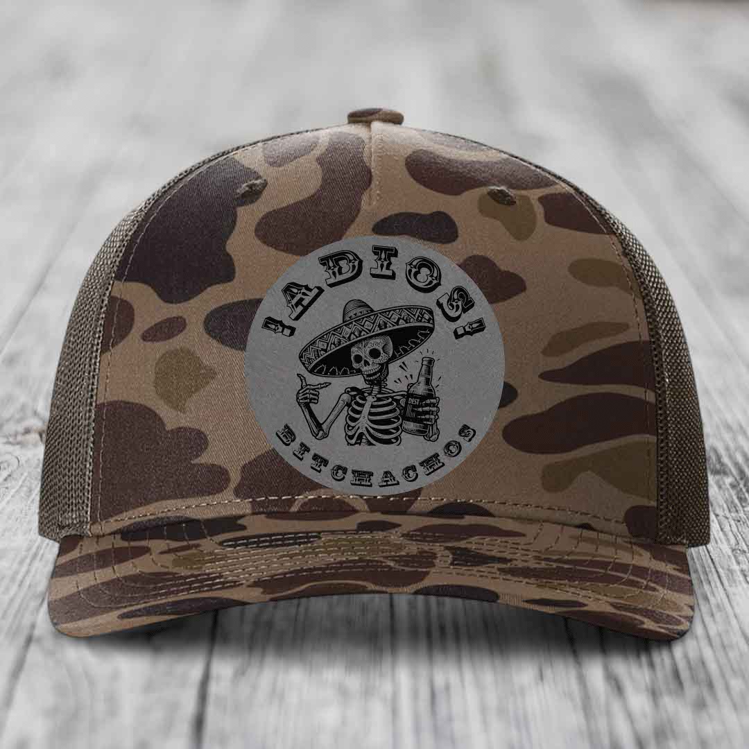 Camouflage trucker hat with a grey circular patch featuring a skeleton in a sombrero holding a beer bottle and the text &