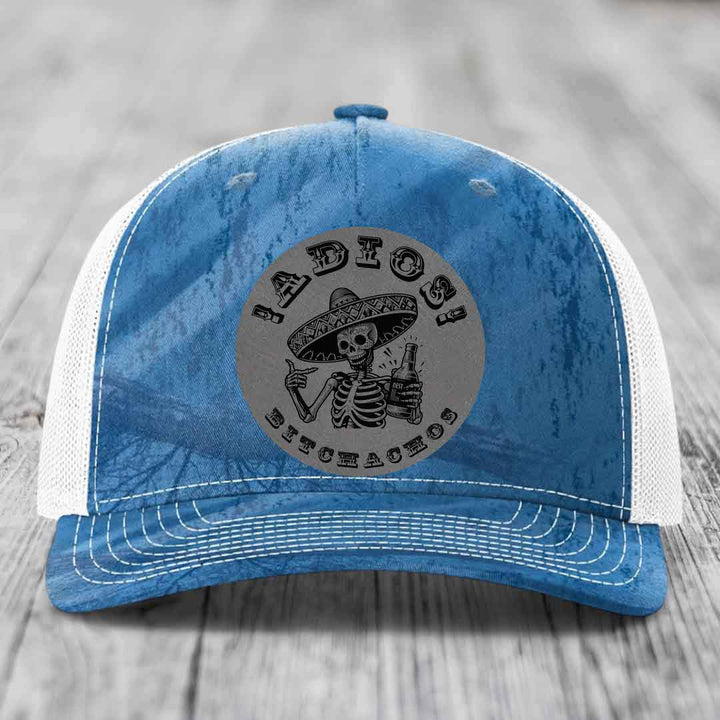 Blue and white trucker hat with a circular grey patch featuring a skeleton in a sombrero holding a beer bottle and the text &