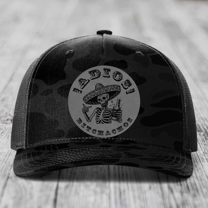 Black camouflage trucker hat with a circular patch featuring a skeleton in a sombrero holding a beer bottle and the text &