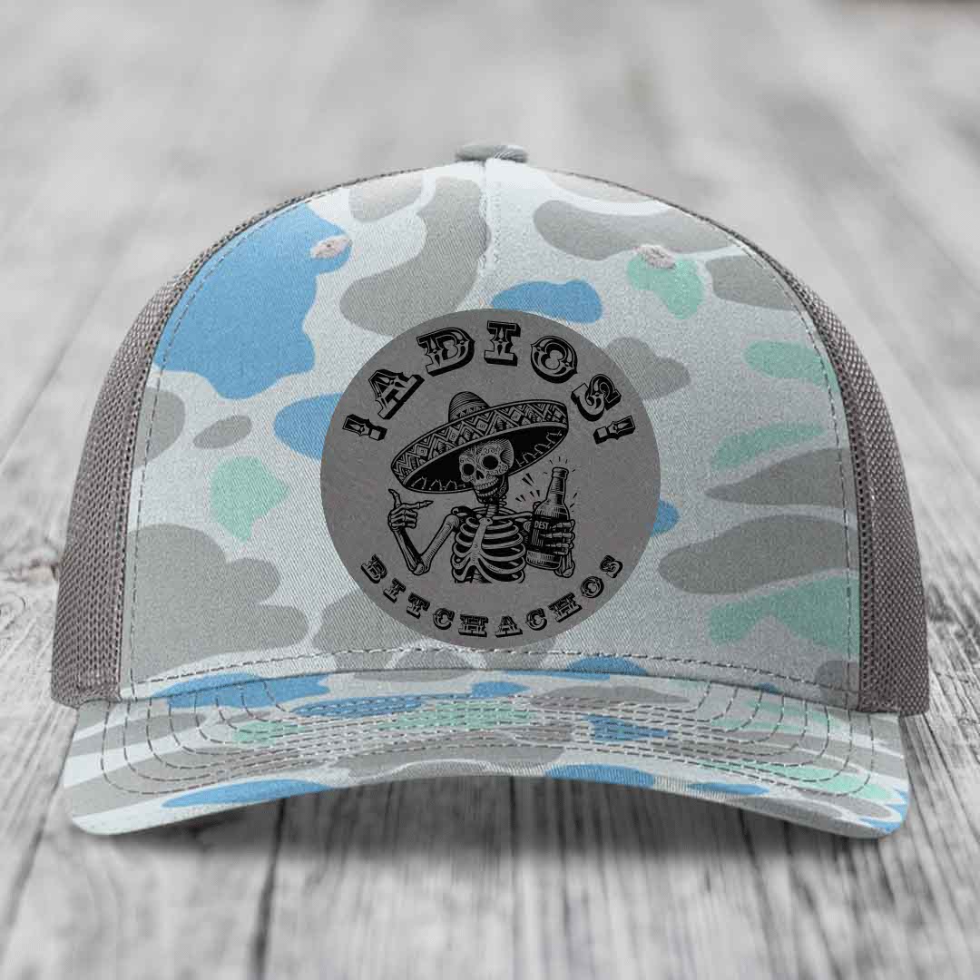 Camouflage trucker hat with grey circular patch featuring skeleton in sombrero holding beer bottle and text &