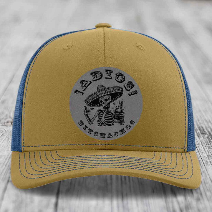 Tan and blue trucker hat with a circular grey patch featuring a skeleton in a sombrero holding a beer bottle and the text &