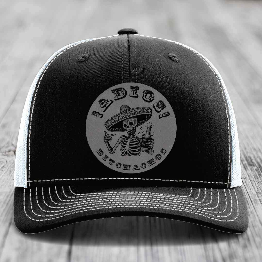 Black trucker hat with white stitching and a grey circular patch featuring a skeleton in a sombrero holding a beer bottle.