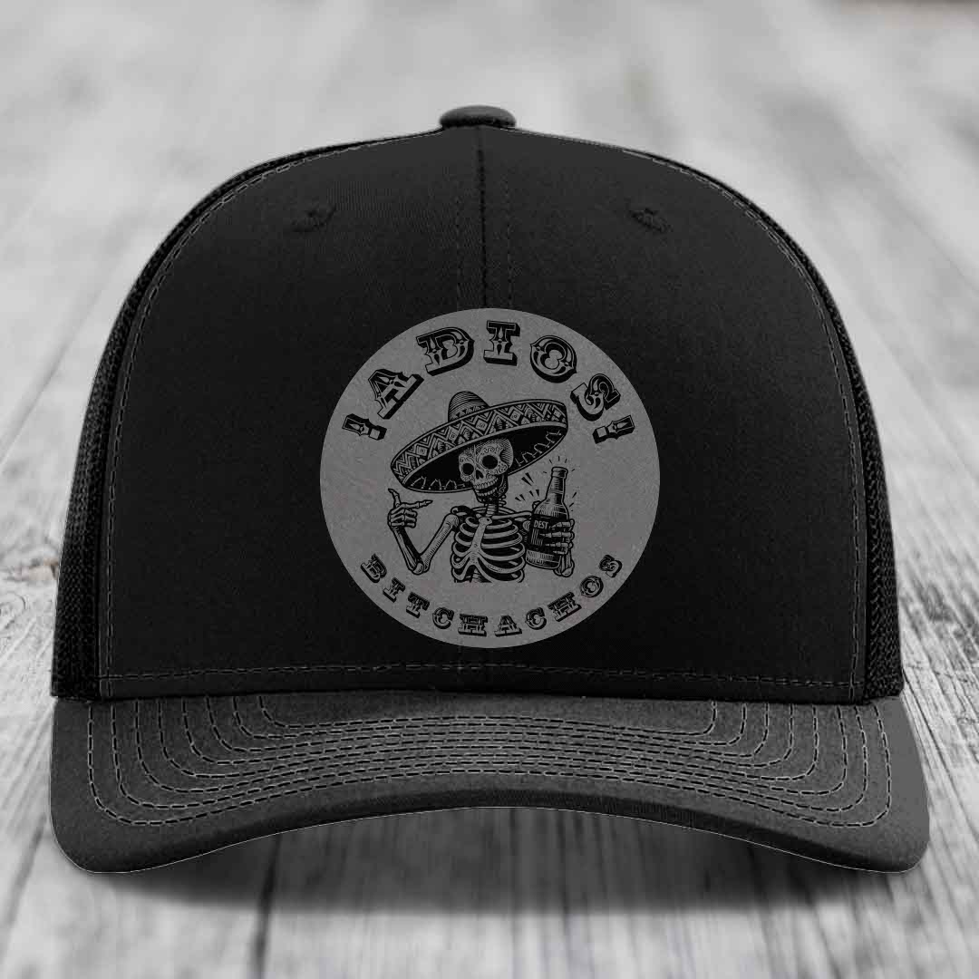 Black trucker hat with a grey circular patch featuring a skeleton in a sombrero holding a beer bottle and the text &