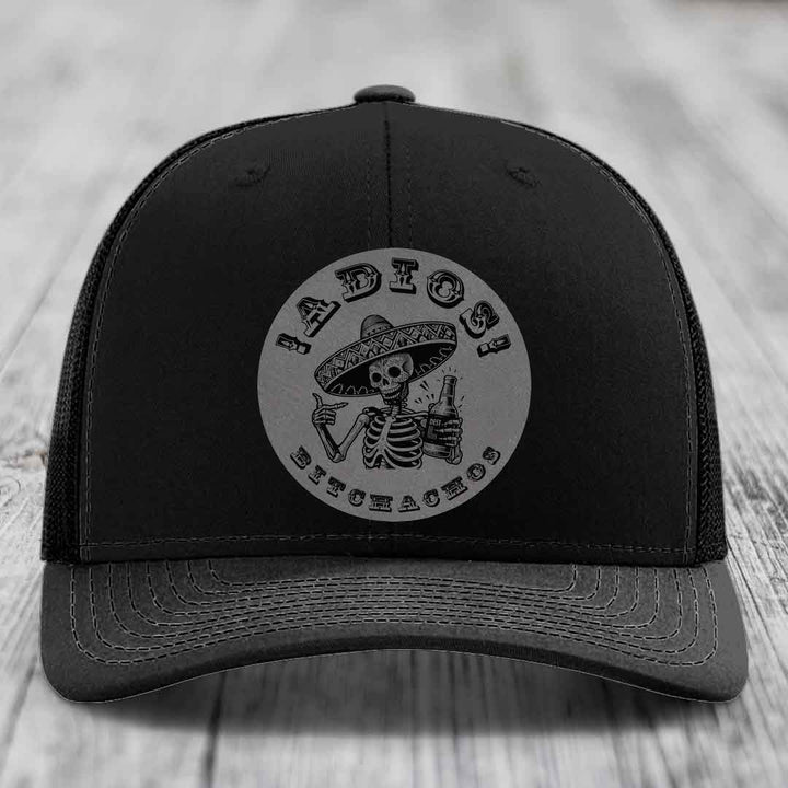 Black trucker hat with a grey circular patch featuring a skeleton in a sombrero holding a beer bottle and the text &