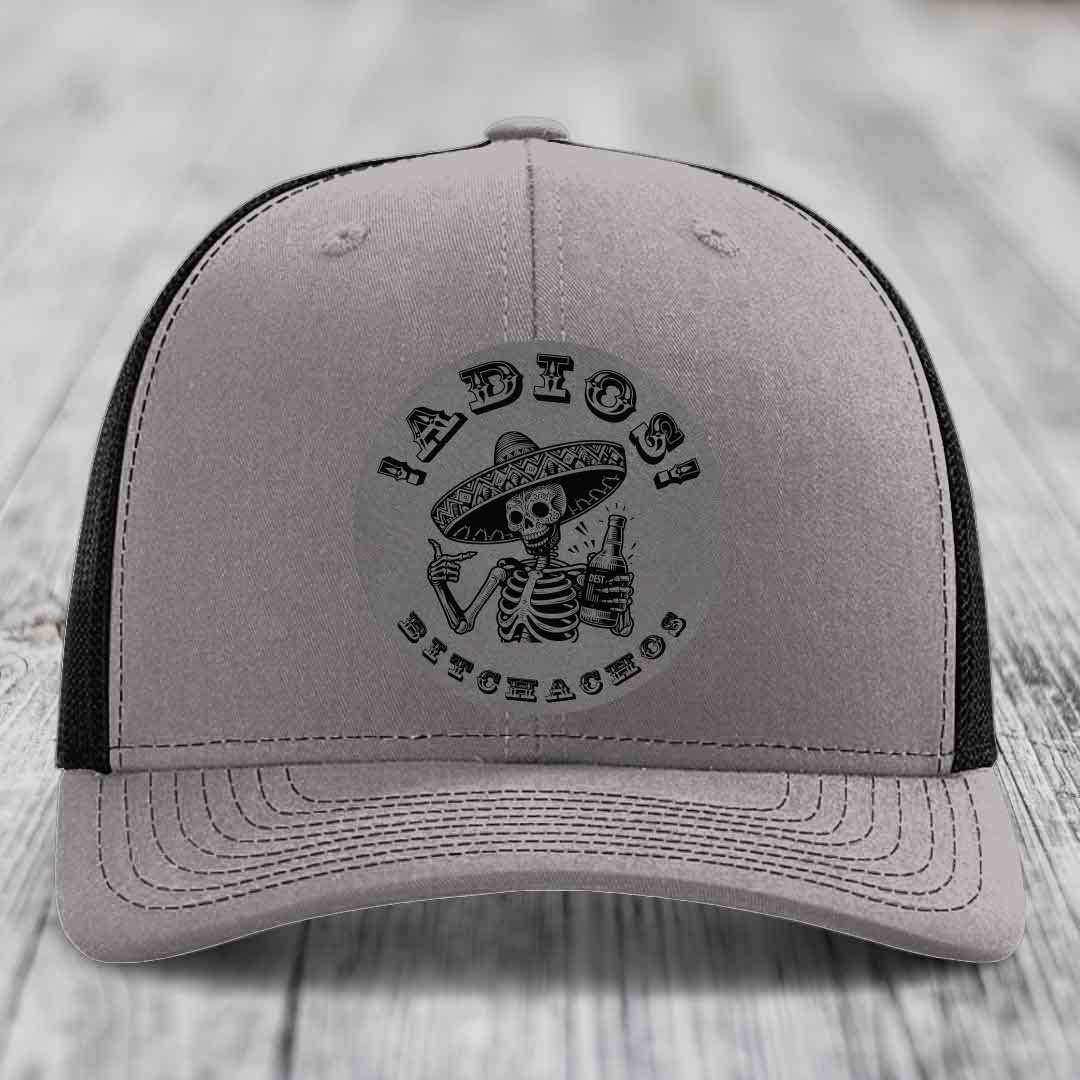 Grey trucker hat with black mesh back and embroidered skull holding bottle patch.