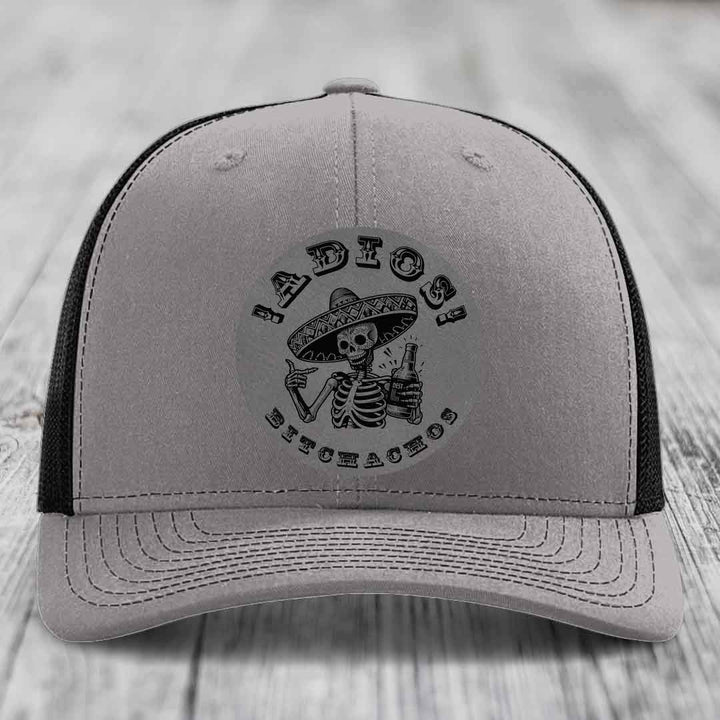 Grey trucker hat with black mesh back and embroidered skull holding bottle patch.