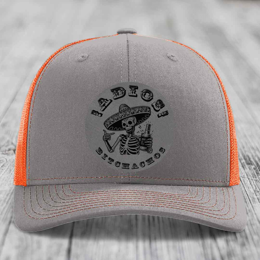 Grey trucker hat with orange mesh back and black skull graphic holding beer bottle under &