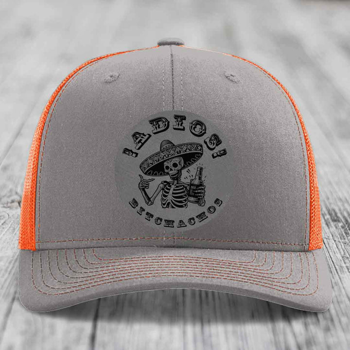 Grey trucker hat with orange mesh back and black skull graphic holding beer bottle under &