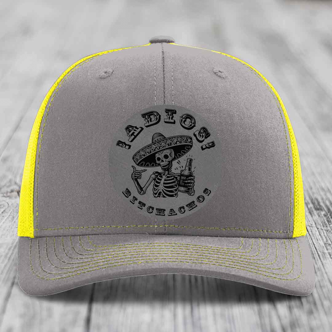 Grey trucker hat with yellow mesh and stitching, featuring a black skull-and-sombrero patch that reads &