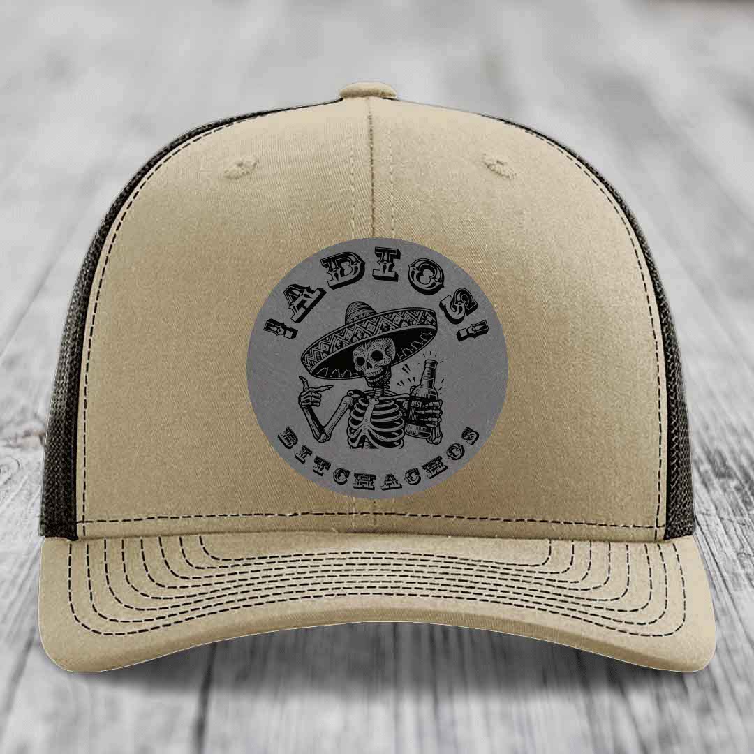 Beige trucker hat with black mesh back and gray circular patch featuring skeleton in sombrero holding beer bottle.