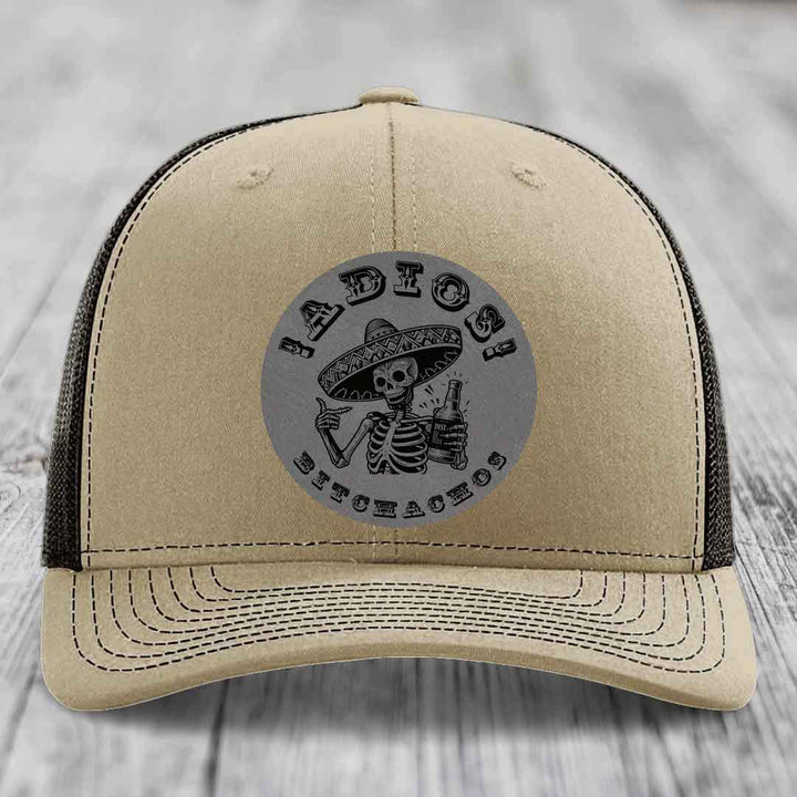 Beige trucker hat with black mesh back and gray circular patch featuring skeleton in sombrero holding beer bottle.