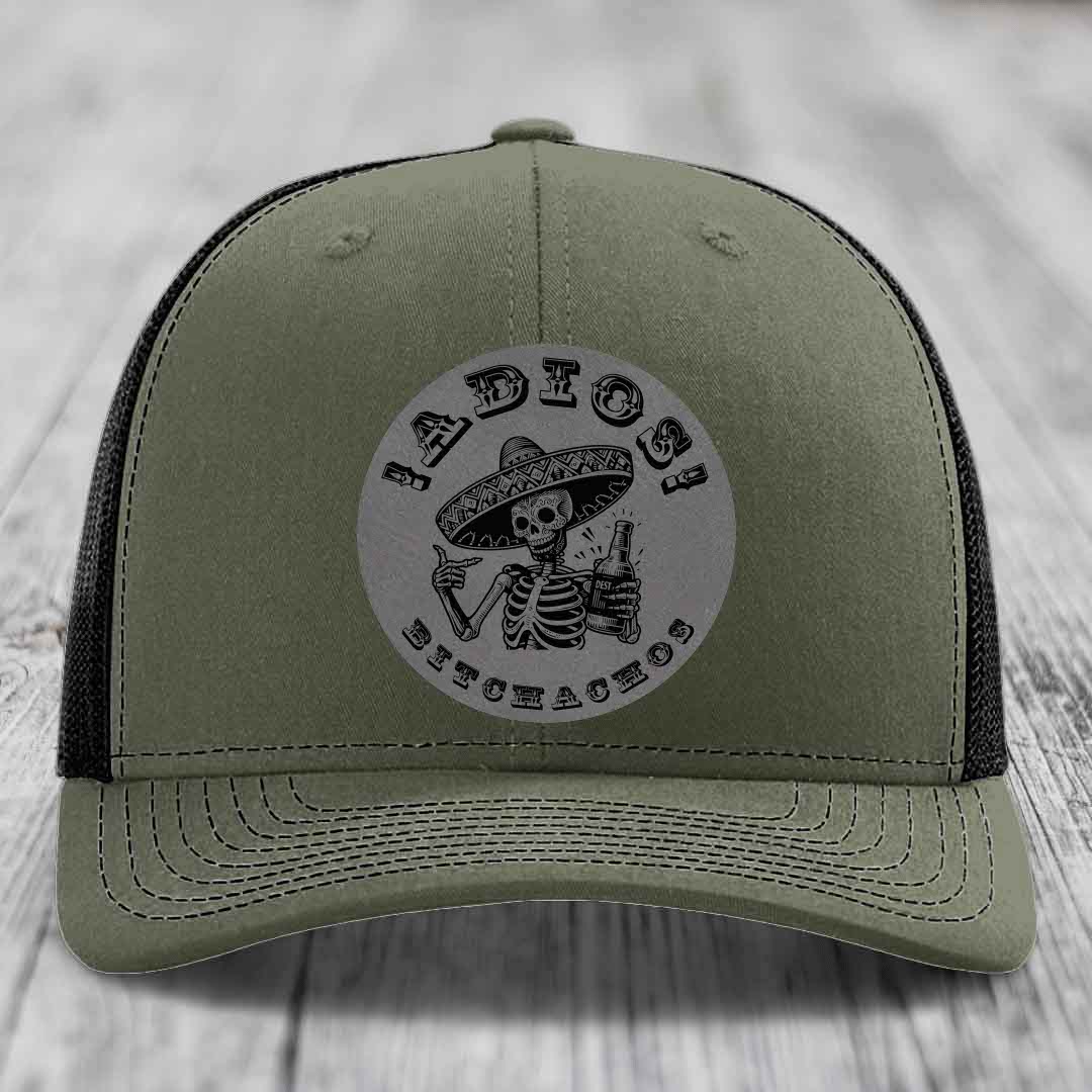 Olive green trucker hat with black mesh back and a circular grey patch featuring a skeleton in a sombrero holding a beer bottle, with the text &