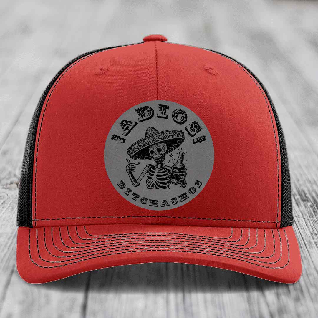 Red trucker hat with black mesh back and gray circular patch featuring a skeleton in a sombrero holding a beer bottle with text &