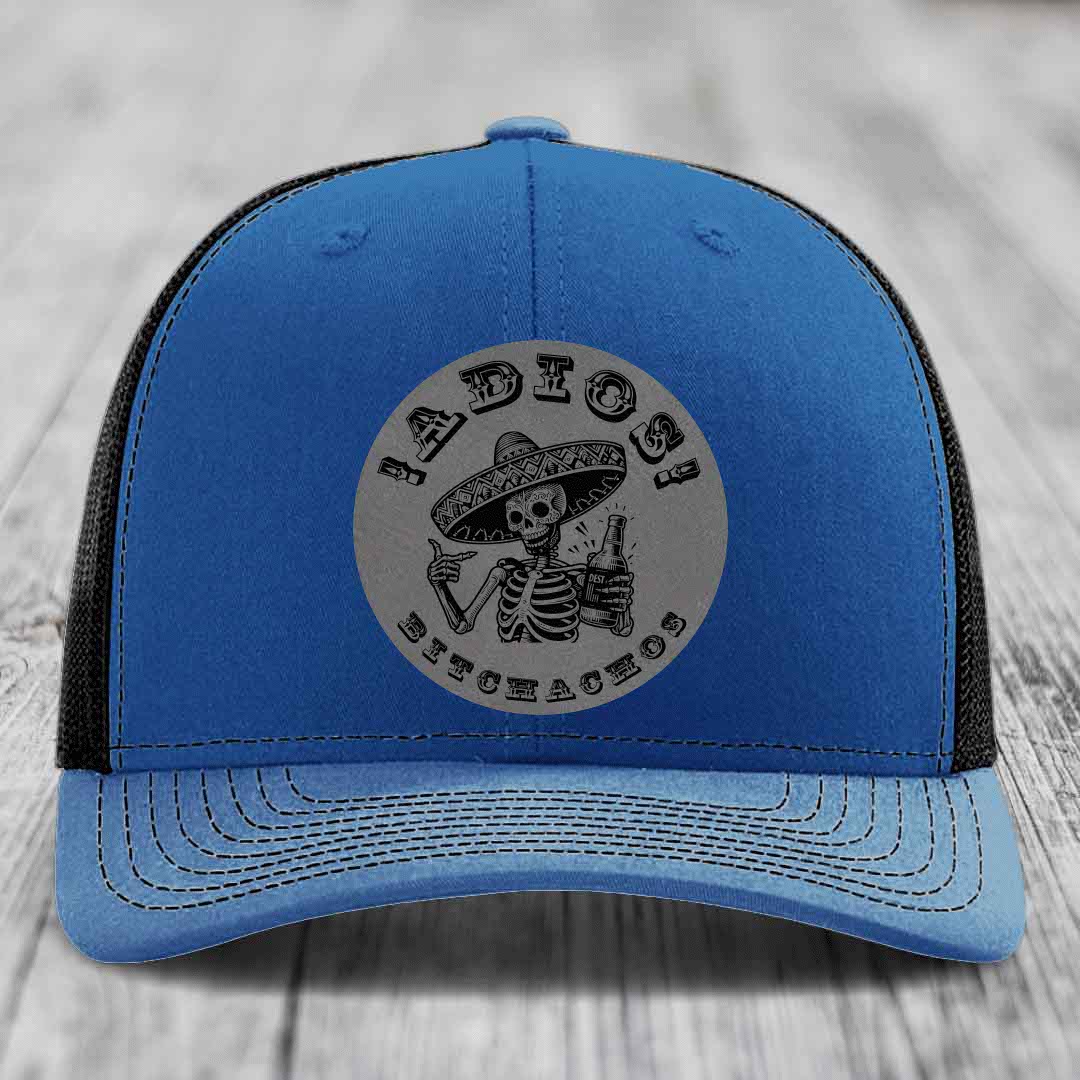 Blue trucker hat with black mesh back and gray circular patch featuring a skeleton in a sombrero holding a beer bottle with text &