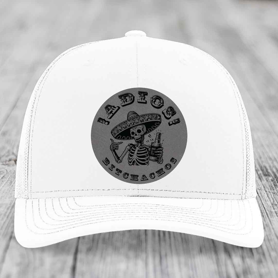 White trucker hat with black circular patch featuring skeleton in sombrero holding beer bottle and text &