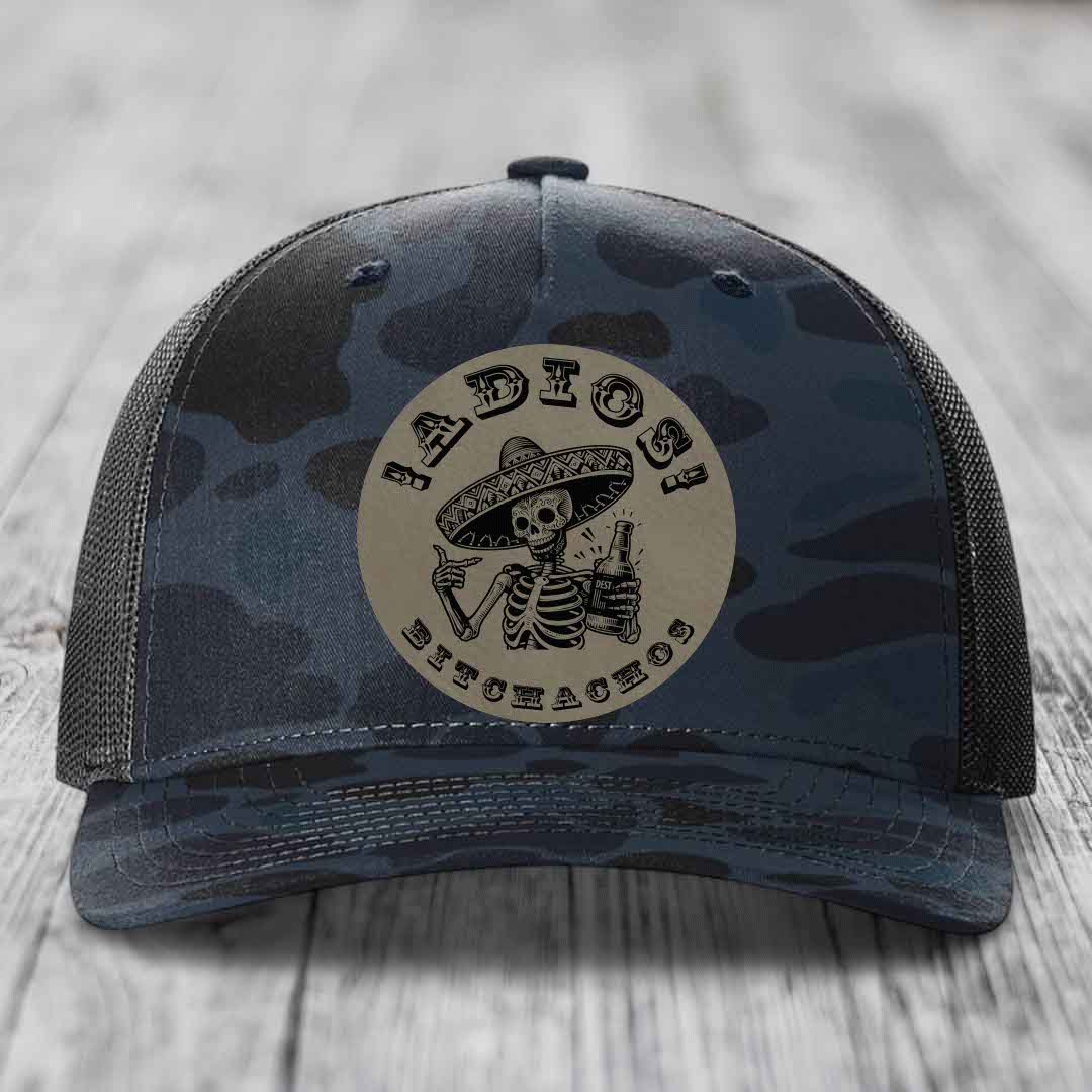 Camouflage trucker hat with a circular patch featuring a skeleton in a sombrero holding a beer bottle and the text &