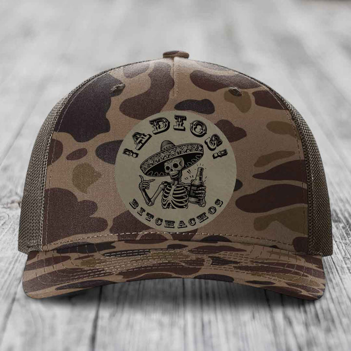 Camouflage trucker hat with a circular patch featuring a skeleton in a sombrero holding a beer bottle and the text &