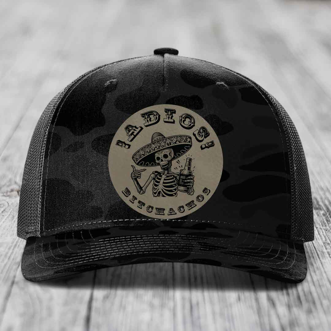 Black camouflage trucker hat with circular patch of skeleton in sombrero holding beer bottle.