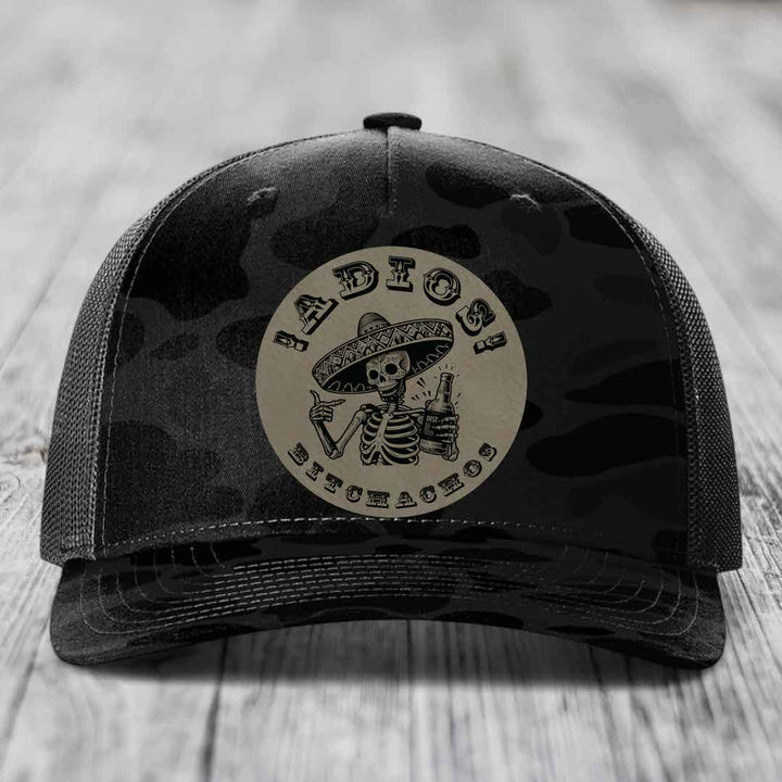 Black camouflage trucker hat with circular patch of skeleton in sombrero holding beer bottle.