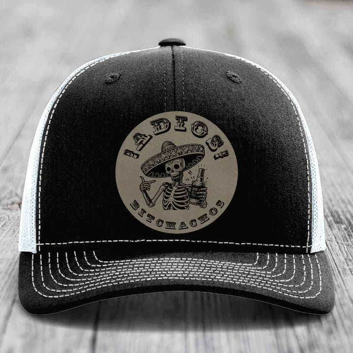 Black trucker hat with white stitching and a circular leather patch featuring a skeleton in a sombrero holding a beer bottle.