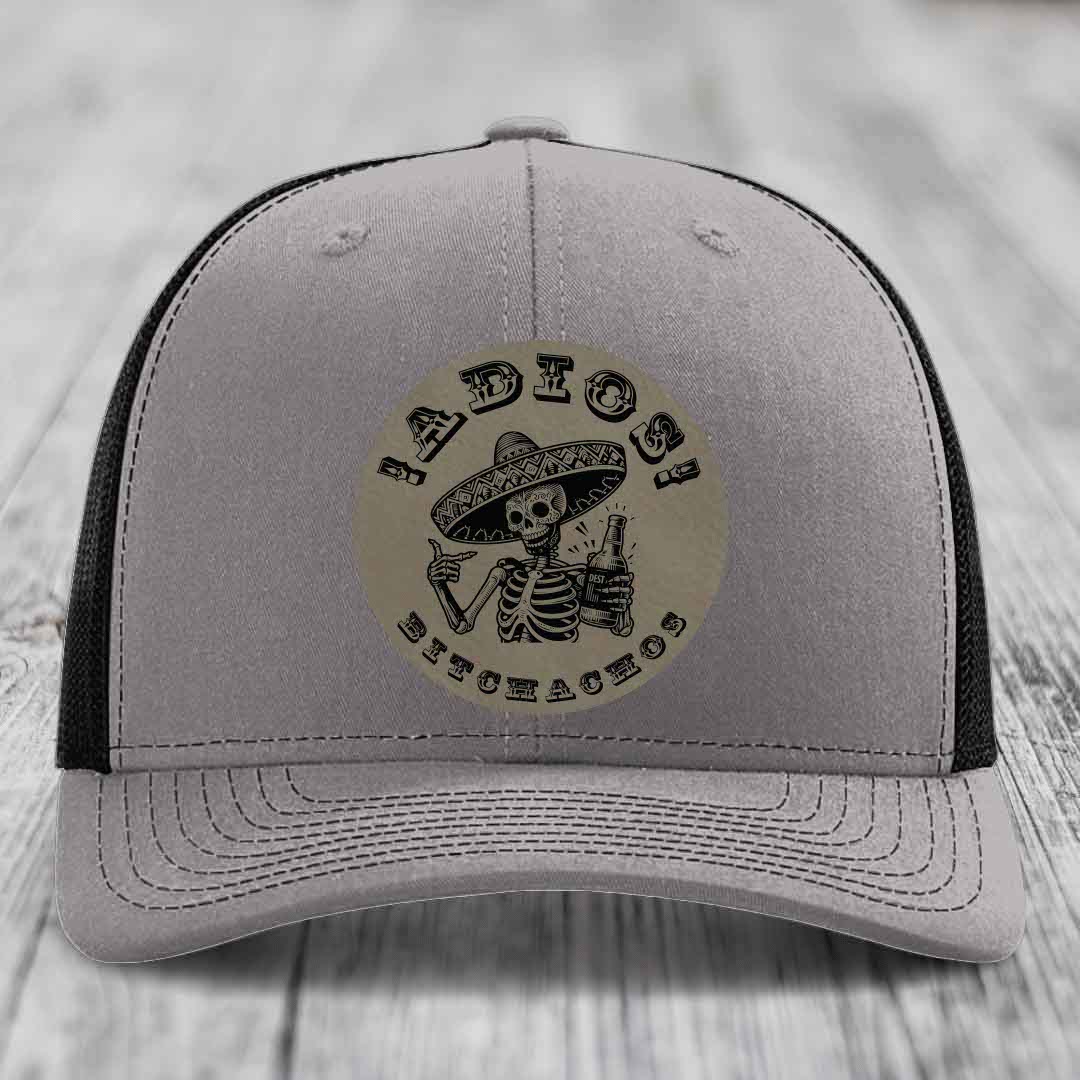 Grey trucker hat with black mesh back and a circular leather patch featuring a skeleton in a sombrero holding a beer bottle.