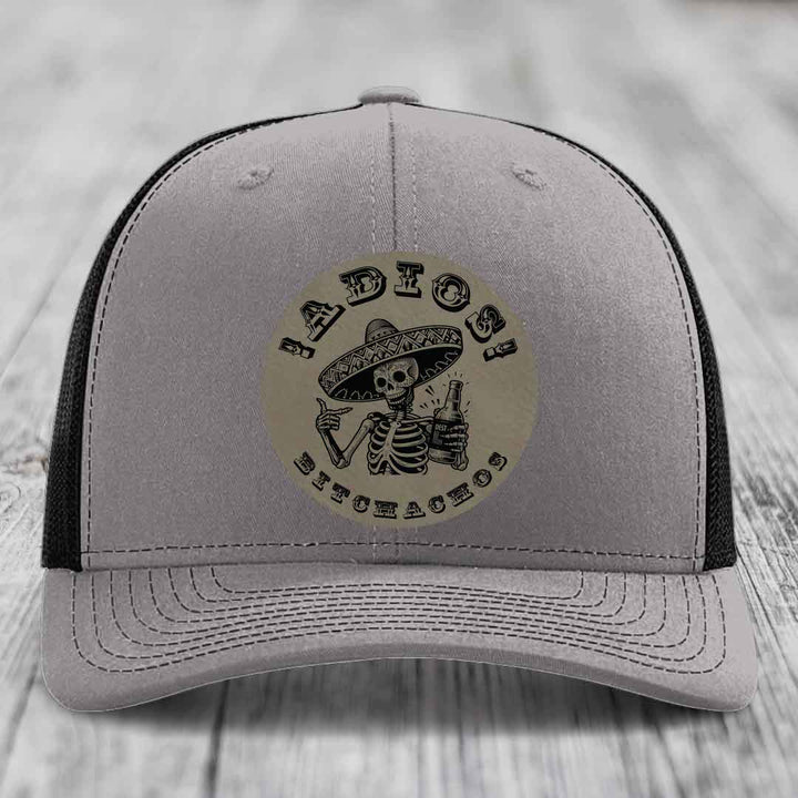 Grey trucker hat with black mesh back and a circular leather patch featuring a skeleton in a sombrero holding a beer bottle.