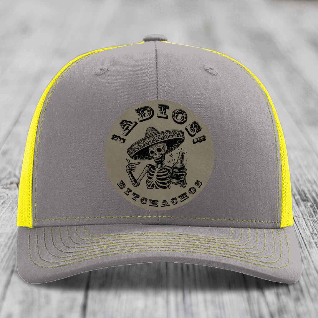 Grey trucker hat with yellow mesh and stitching, featuring a circular patch of a skeleton in a sombrero holding a beer bottle with &