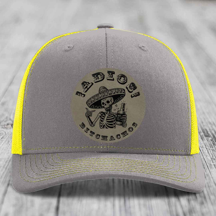 Grey trucker hat with yellow mesh and stitching, featuring a circular patch of a skeleton in a sombrero holding a beer bottle with &